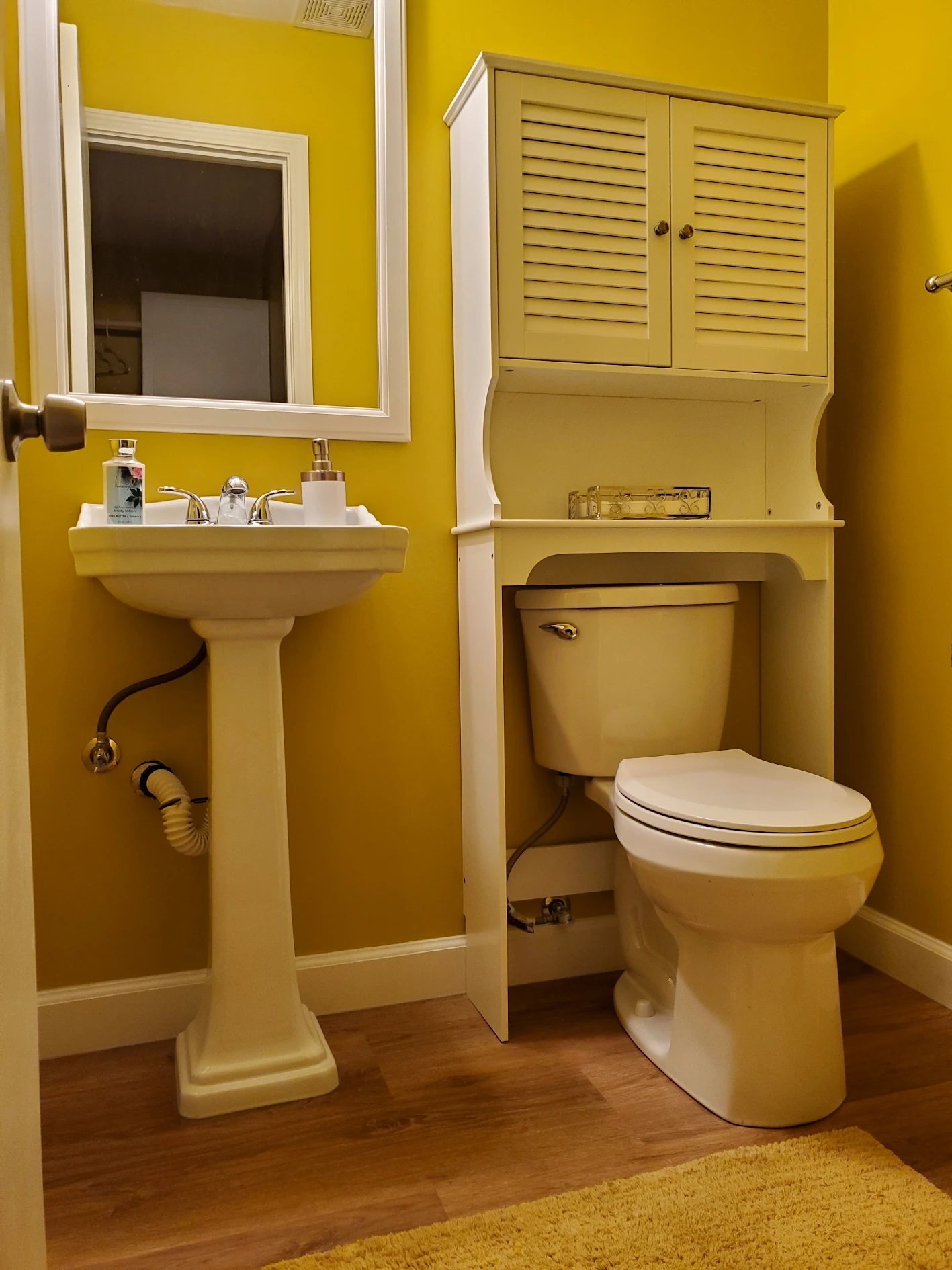 Half bath painted deep yellow with a pedestal sink, framed mirror, and white over-toilet storage cabinet